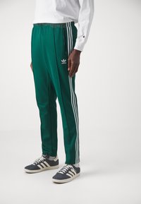 Gröna Adidas-joggingbyxor med vita sidskärningar, elastisk midja och en liten logotyp på vänster lår, tillsammans med mörka sneakers.