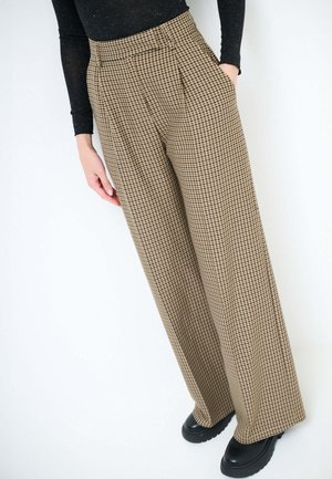 Pantaloni a gamba larga realizzati in tessuto pied de poule nei toni del beige e marrone. Caratterizzati da vita alta, pieghe, tasche e abbinati a scarpe platform nere.