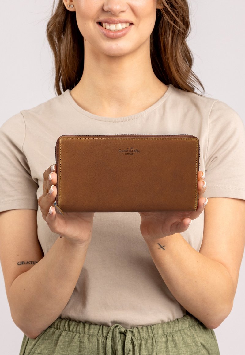 Brown leather wallet with a zip closure, features a smooth texture and embossed logo on the front. Held in hands against a neutral background.