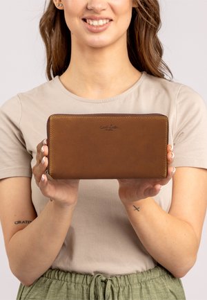 Cartera de cuero marrón con cierre de cremallera, presenta una textura suave y un logo en relieve en la parte frontal. Sostenida en las manos contra un fondo neutro.