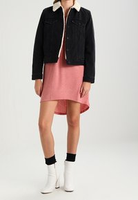 Veste en jean noire avec un col en fausse fourrure crème, portée sur une robe rose. Le mannequin porte des bottines blanches et des chaussettes noires, illustrant un look décontracté.