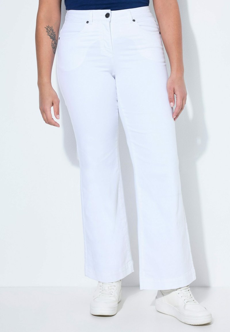 Femme portant un pantalon large blanc taille haute avec fermeture à boutons et zip, assorti à des baskets blanches et un haut bleu marine. Tatouage visible sur le bras.