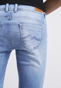 Světle modré džíny s úzkým střihem, s zadními kapsami a hnědou koženou nášivkou. Výrazné švy na okrajích kapes.