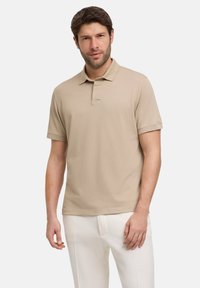 Polo shirt color beige chiaro con colletto, maniche corte e patta con bottoni; indossato con pantaloni bianchi. Texture liscia, design informale.