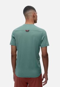 Zaļa sporta krekls ar īsām piedurknēm, gluda tekstūra, noapaļota kakla izgriezums un neliela sarkana akcenta detaļa aizmugurē.