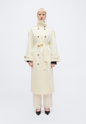 Άτομο με κοντά ξανθά μαλλιά φοράει μακρύ κρεμ παλτό διπλής όψης με ψηλό γιακά, ζώνη στη μέση, φαρδιά μανίκια και μαύρα μυτερά παπούτσια.