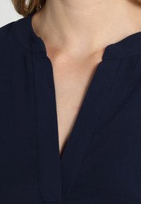 Haut bleu marine avec un décolleté en V, fabriqué dans un tissu lisse. Présente un design épuré et des détails de couture subtils près du décolleté.