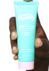 Turkusowa tubka ze ściskaną nakrętką zawierająca matujący krem, z różową podstawą. Na przedniej stronie znajduje się tekst "Merci Handy" oraz składniki. Pojemność 50 ml.