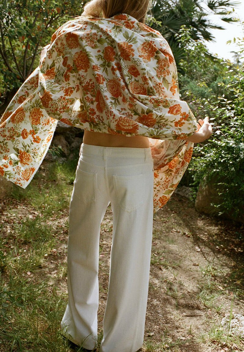 Personne à l'extérieur drapant un châle à motifs floraux orange et vert sur ses épaules, portant un pantalon blanc à jambes larges sur un chemin de terre.