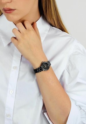 Sort metalur med rund kasse og tekstureret sort urskive. Har en ledet rem og en krystalbesat bezel. Båret på en lys hånd.