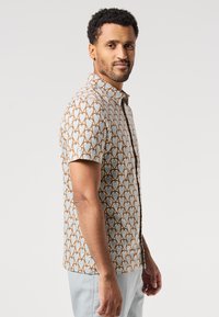 Homme avec des cheveux courts et bouclés portant une chemise à manches courtes avec des motifs de cœurs bleus et bruns, se tenant de profil contre un fond uni.