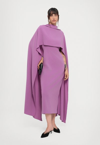 Lilla kaftanikleid, millel on kõrge kaelus, keha kallistav siluett ja voolav kaftanikate detail. Kombineeritud mustade teravate kontsade ja käekotiga.