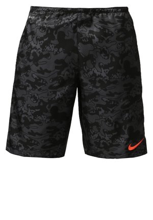 Mies kävelee päällään kirkkaan oranssi Nike-urheilupaita, mustat maastokuvioiset shortsit, valkoiset raidalliset sukat ja mustat urheilukengät.