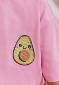 Rosa t-shirt med en strukturerad, broderad avokado-design med ett leende ansikte, en rund brun kärna och svart kontur på vänster sida.