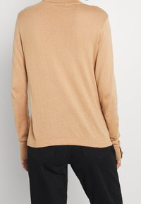 Maglione beige a maniche lunghe con finitura testurizzata, dotato di colletto e polsini a coste, abbinato a pantaloni neri.