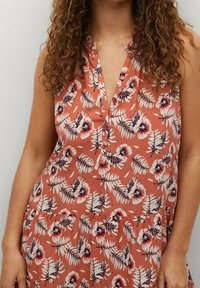 Robe sans manches à motifs floraux couleur corail avec un décolleté en V boutonné, présentant des motifs de fleurs blanches et bleu foncé, fabriquée dans un textile doux et fluide.