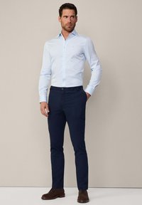 Lichtblauw gestreefd overhemd met lange mouwen, gecombineerd met marineblauwe broek met zijzakken en bruine schoenen.