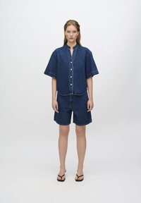 Outfit in denim con una camicia a maniche lunghe e colletto, abbinata a pantaloni corti. Colore blu scuro, bordi sfrangiati e dettagli di cucitura a contrasto.