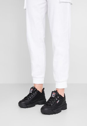 Ben iklædt hvide joggerbukser og sorte FILA chunky sneakers med tykke såler og logodetaljer på tunger og sider.