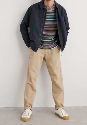 Homme portant un pull rayé, une veste zippée bleu marine, un pantalon beige et des baskets blanches, debout contre un mur uni, les mains dans les poches.