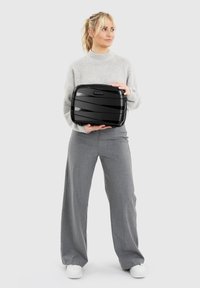 Vrouw in grijze trui en broek die een zwarte harde koffer met het "REDOLZ" logo vasthoudt, tegen een effen lichte achtergrond.