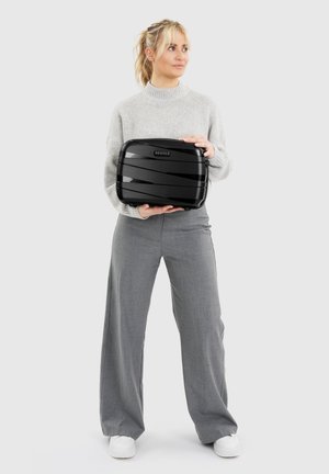 Vrouw in grijze trui en broek die een zwarte harde koffer met het "REDOLZ" logo vasthoudt, tegen een effen lichte achtergrond.