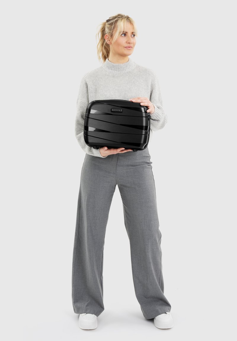 Vrouw in grijze trui en broek die een zwarte harde koffer met het "REDOLZ" logo vasthoudt, tegen een effen lichte achtergrond.