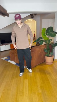 Pullover marrone, pantaloni neri e sneakers argento. Il pullover presenta un colletto e un piccolo logo. Nel fondo ci sono una pianta e un divano.