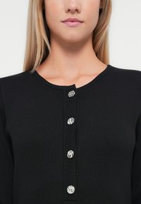 Μαύρο ριγέ πουλόβερ με στρογγυλή λαιμόκοψη και πέντε διακοσμητικά κουμπιά με αντίθετα σχέδια κατά μήκος της πλαινής ραφής.