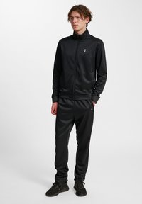 Sort atletisk træningsdragt lavet af glat, letvægtsstof, der har en lynlåsjakke og tætsiddende bukser, prydet med et lille logo.