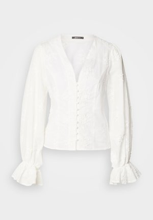 Blouse blanche à manches longues avec col en V, boutons recouverts de tissu sur le devant, broderie florale et poignets volantés sur un fond uni.