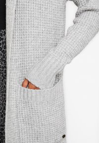 Gray knitted cardigan featuring a textured surface, ribbed cuffs, and front pockets. Fabric shows a soft, cozy appearance.