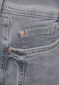 Ljust grå denimjeans har en synlig ficka med kopparnitar och kontrasterande bruna sömmar. Det texturerade tyget antyder hållbarhet.