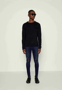 Homem que usa uma camisa preta de mangas compridas, jeans skinny escuros rasgados, sapatos pretos e óculos de sol, em pé contra um fundo claro e liso.