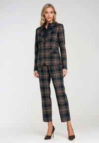 Garnitur w kratę w ciemnozielonych, brązowych i beżowych odcieniach. Dopasowana marynarka z klapami, pasujące wąskie spodnie i błyszczące, ciemne obcasy.
