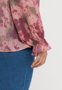 Main reposant à côté d'un jean bleu, portant un chemisier rose à fleurs avec un poignet froncé et volanté sur la manche, sur fond blanc.
