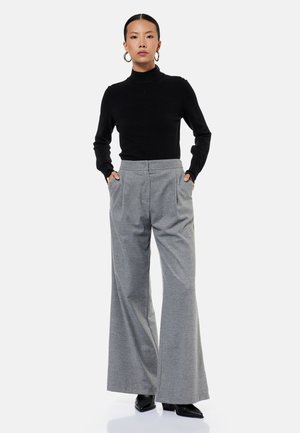 Nainen pukeutunut mustaan poolokaulaiseen neulepaitaan, harmaisiin leveälahkeisiin housuihin, mustiin kärkikorkokenkiin ja renkaanmuotoisiin korvakoruihin, seisoo kädet taskuissa.