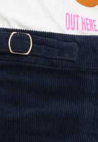 Witte shirt met roze "OUT HERE" tekst, ingetikt in marineblauwe corduroy broek met metalen sluiting op de taille.