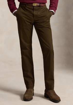 Homme portant un pantalon vert olive foncé, des bottes en daim marron, une ceinture marron et une chemise à manches longues bordeaux, les mains dans les poches.