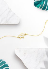 Guld elefantberlockarmband med ett delikat kedja, placerat mot en vit marmorbakgrund och gröna tropiska blad.