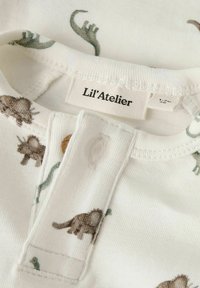 Wit katoenen babyshirt met bruine en groene dinosaurusprints. Voorzien van een knoopsluiting en een label met de tekst "Li'l' Atelier."