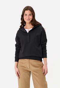 Schwarzer Kapuzenpullover mit Reißverschluss am Hals, ausgestattet mit gerippten Bündchen, lockerer Passform und glatter Textur, kombiniert mit beigen Hosen.