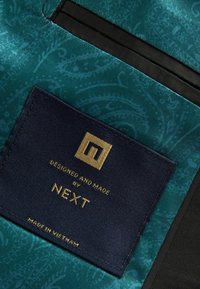 Tissu sarcelle avec un motif subtil, comportant une étiquette marine avec un texte doré, indiquant "Conçu et fabriqué par NEXT, fabriqué au Vietnam."