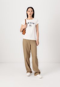 T-shirt bianca con grafica "NEW DENIM", pantaloni beige a gamba larga e sneakers bianchi. Borsa marrone con tracolla corta. Texture lisce.