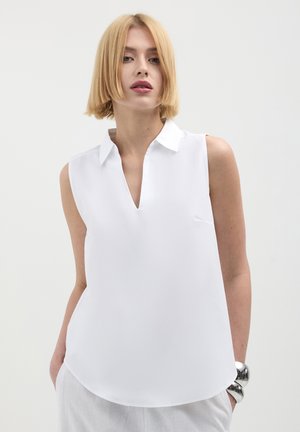 Donna bionda con capelli corti, che indossa una blusa bianca senza maniche con colletto, abbinata a pantaloni di colore chiaro e un grande bracciale d'argento.