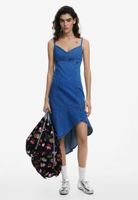 Denimmekko, jossa on muotoiltu yläosa ja epäsymmetrinen helma, säädettävillä olkaimilla ja sinisillä sävyillä. Musta laukku, jossa on kukkakuosi. Hopeiset kengät.