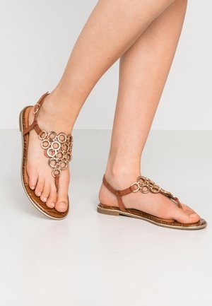 Pieds portant des sandales plates marron avec des détails de chaîne circulaire dorée sur fond clair.