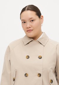 Beige trenchcoat med dobbeltradet design, med store mørke knapper og sidelommer. Laget av glatt, lett stoff.