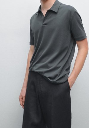 Homme portant un polo gris foncé à manches courtes légèrement rentré dans un pantalon noir ample, avec une main dans la poche, debout devant un fond clair.