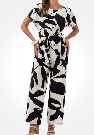 Kobieta ubrana w kombinezon w czarno-biały abstrakcyjny wzór liści, z krótkimi rękawami, wiązany w talii, oraz beżowe sandały na szerokim obcasie.